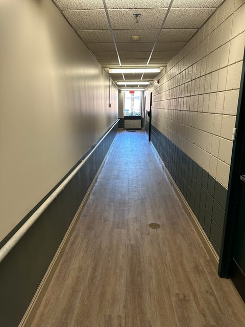 Long narrow hallway with beige walls, white ceiling tiles, fluorescent lighting, wood flooring, and white baseboards receding into the distance
