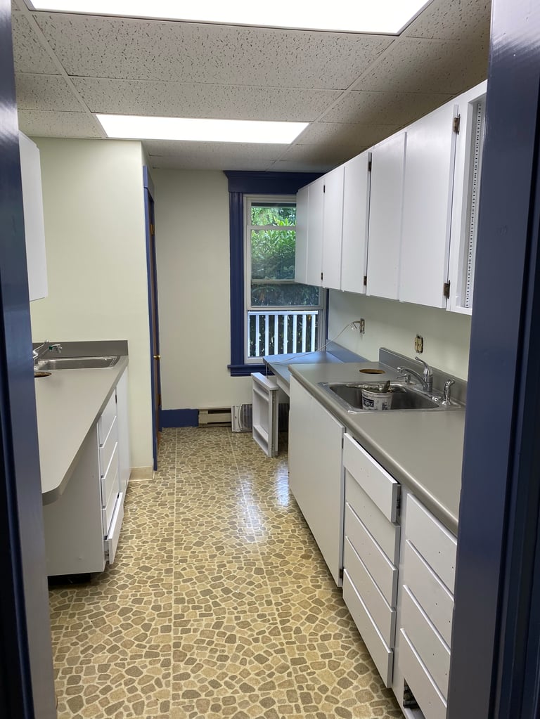Narrow galley kitchen with white cabinets, stainless steel sink, and beige tile flooring, featuring blue walls and a window with white railings